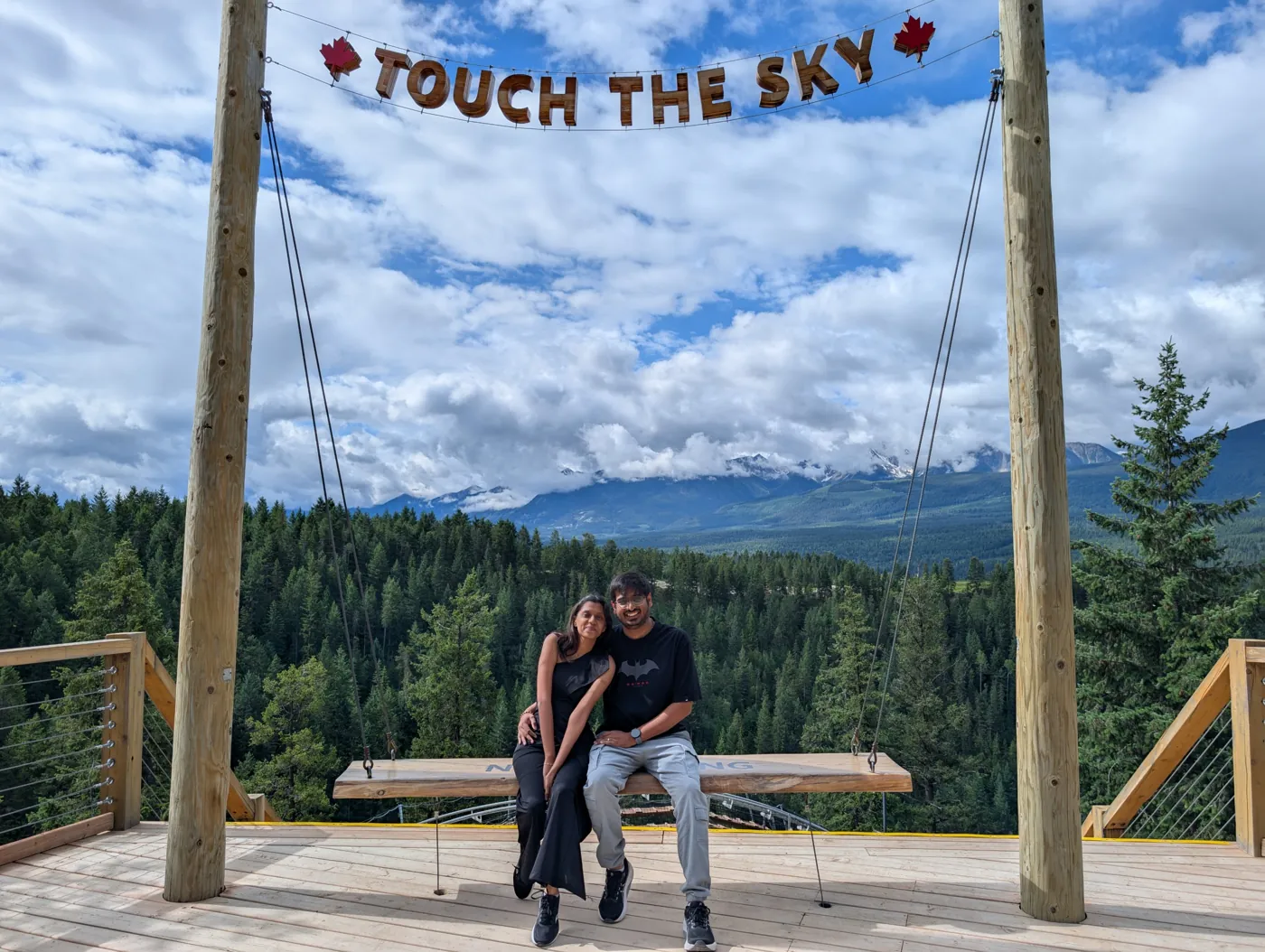 Kishan and Jinal sitting together on a swing under a Touch the Sky sign, with forest and mountains behind them